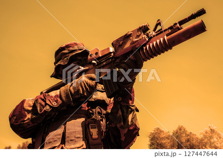 Soldier in camouflage attire and field helmet aiming a rifle with mounted optic under a yellow-orange sky, with trees in the background. 127447644