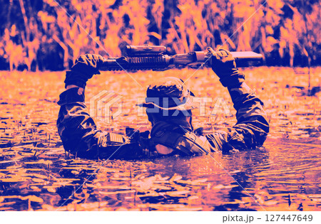 A soldier wearing camouflage holds a firearm above his head while wading through water surrounded by reeds in a dramatic color filter. 127447649