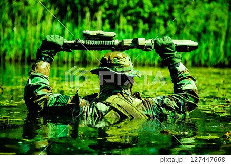 Soldier in camouflage holding a rifle above water while wading through a green swampy area surrounded by vegetation. Soldier in camouflage holding a rifle above water while wading through a green swampy area surrounded by vegetation. 127447668