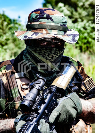 Soldier with camo gear and face covering, holding a rifle with optics in a green, wooded outdoor setting under clear blue sky. Soldier with camo gear and face covering, holding a rifle with optics in a green, wooded outdoor setting under clear blue sky. 127447671