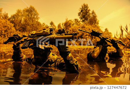 Two camouflaged soldiers wade through water holding rifles above their heads, with trees in the background on a clear day. Two camouflaged soldiers wade through water holding rifles above their heads, with trees in the background on a clear day. 127447672