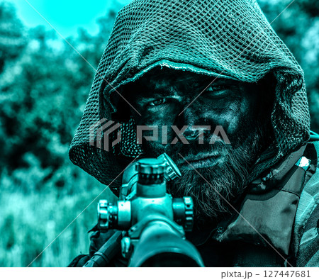 Intense sniper with a hood and camouflage face paint aiming a rifle, set against a blurred natural background with a blue color tone. Intense sniper with a hood and camouflage face paint aiming a rifle, set against a blurred natural background with a blue color tone. 127447681