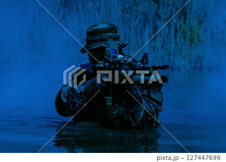 Soldier in camouflage gear wading in water, holding a rifle with tactical equipment, surrounded by dense vegetation under a blue-tinted light. 127447699