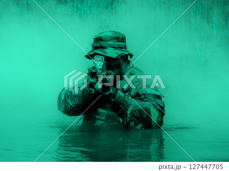 Soldier in a camouflage uniform and hat aiming a rifle while half-submerged in water, with a background of green mist. Soldier in a camouflage uniform and hat aiming a rifle while half-submerged in water, with a background of green mist. 127447705