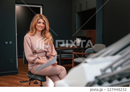 Sitting and holding document. Beautiful young female doctor is indoors in the cabinet 127448232