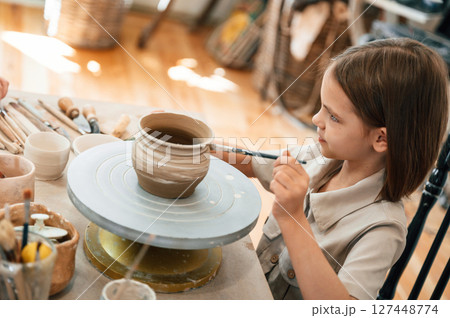 Painting the pot by brush. Little girl is learning how to do pottery in the workshop 127448774