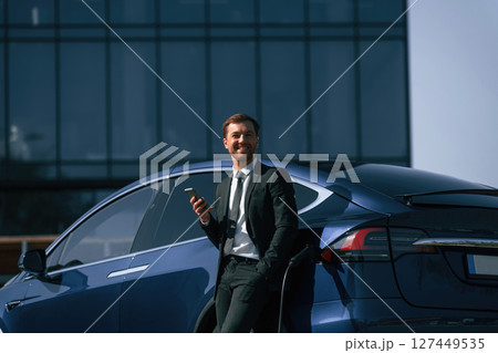 Smiling, holding smartphone. Businessman is standing near his electric car outdoors 127449535