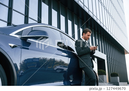 Holding smartphone. Businessman is standing near his car outdoors Holding smartphone. Businessman is standing near his car outdoors 127449670
