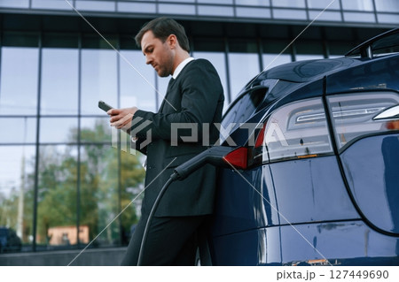 Waiting for a charge. Businessman is standing near his electric car outdoors Waiting for a charge. Businessman is standing near his electric car outdoors 127449690