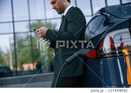 Electric car is charging, businessman with smartphone is waiting Electric car is charging, businessman with smartphone is waiting 127449692