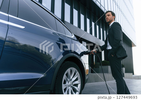 Man in suit and tie is holding charger and standing near his electric car outdoors Man in suit and tie is holding charger and standing near his electric car outdoors 127449693