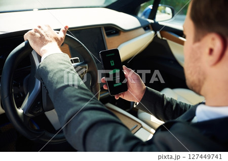 Holding smartphone with charging progress app. Businessman in suit is driving his electric car Holding smartphone with charging progress app. Businessman in suit is driving his electric car 127449741