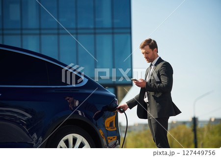 Holding the charger. Businessman is standing near his electric car outdoors Holding the charger. Businessman is standing near his electric car outdoors 127449756