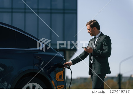 Holding the charger. Businessman is standing near his electric car outdoors 127449757