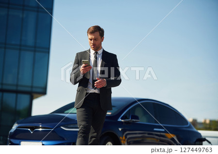 Serious facial expression. Businessman is standing near his electric car outdoors Serious facial expression. Businessman is standing near his electric car outdoors 127449763