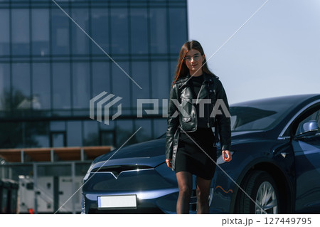 In black jacket and skirt. Young woman is near her electric car outdoors 127449795