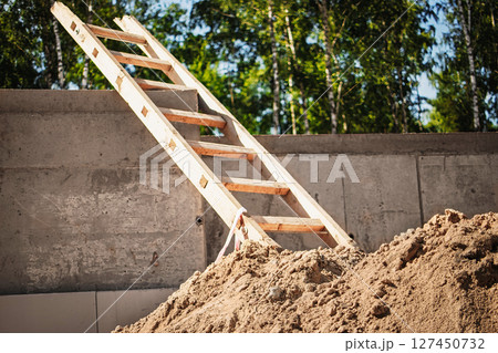 Construction site with wooden ladder propped against a concrete wall and a mound of sand in a wooded area Construction site with wooden ladder propped against a concrete wall and a mound of sand in a wooded area 127450732