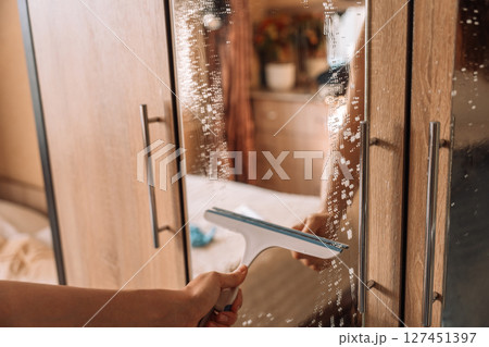 Cleaning a wardrobe mirror with a rubber squeegee, removing soap and water during routine household chores. Cleaning a wardrobe mirror with a rubber squeegee, removing soap and water during routine household chores. 127451397