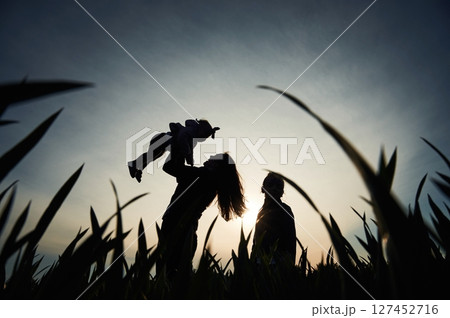 Silhouette of woman that holding her daughter in hands. Another girl is standing and watching 127452716