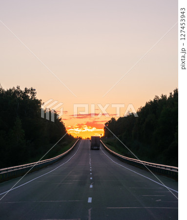 Sunset on an open highway with a lone truck traveling in the distance against a colorful sky Sunset on an open highway with a lone truck traveling in the distance against a colorful sky 127453943