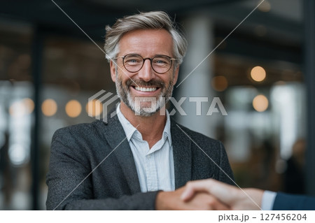 A confident man with gray hair, glasses, and a beard smiles broadly while shaking hands with another individual. The background features a stylish office with bright lighting and blurred figures. 127456204