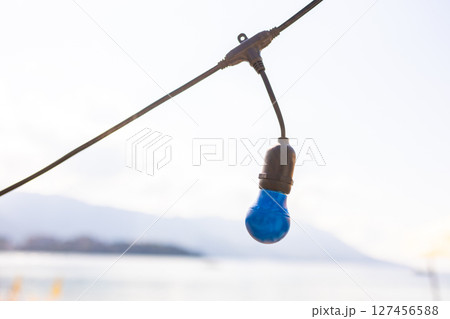 Blue light bulb hanging above beach sand. Decorative lighting element with blurred seaside background and wooden terrace. Blue light bulb hanging above beach sand. Decorative lighting element with blurred seaside background and wooden terrace. 127456588