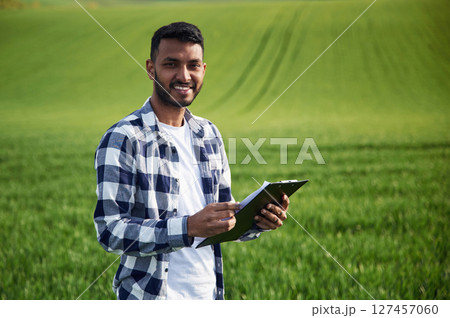 Standing and holding notepad. Handsome Indian man is on the agricultural field 127457060