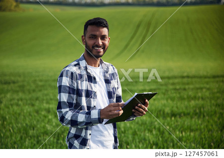 Standing and holding notepad. Handsome Indian man is on the agricultural field Standing and holding notepad. Handsome Indian man is on the agricultural field 127457061