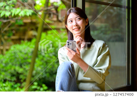 スマホ片手に外の景色を見つめる若い女性のやさしいまなざしと日常の一幕 スマホ片手に外の景色を見つめる若い女性のやさしいまなざしと日常の一幕 127457400