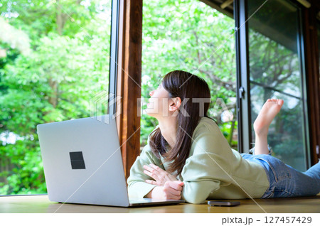窓辺に体を向けてノートパソコンを見上げる女性が一息ついて周囲の景色に目をやる 127457429