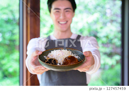 エプロン姿の男性が笑顔でハンバーグを差し出しカメラに向かって料理をアピールしている 127457459
