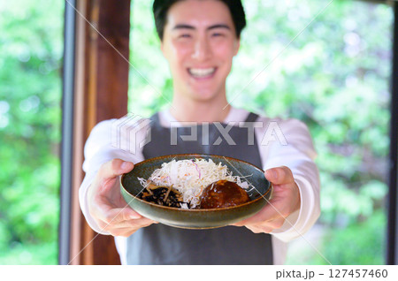 カメラ前方にハンバーグを両手で差し出すエプロン姿の男性が満面の笑顔を見せている カメラ前方にハンバーグを両手で差し出すエプロン姿の男性が満面の笑顔を見せている 127457460