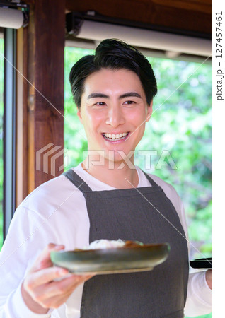 料理を手に持ち柔らかく微笑むエプロン姿の男性が自然光の中でリラックスして立つ 127457461