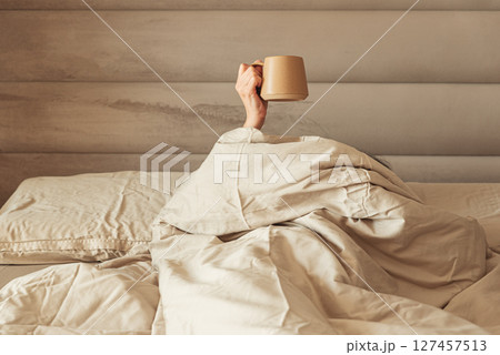 A hand holding a coffee mug emerging from a cozy bed. 127457513