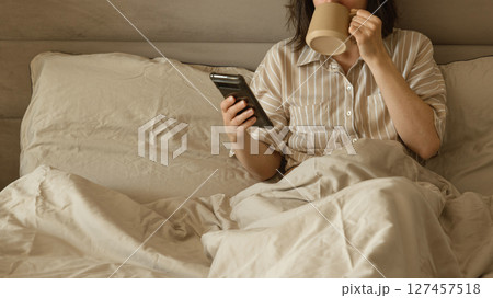 Woman drinking coffee and using phone in bed in the morning. Woman drinking coffee and using phone in bed in the morning. 127457518