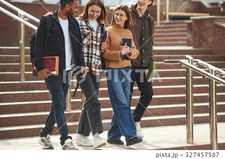 Walking, holding notepads. Four young students in casual clothes are together outdoors 127457587