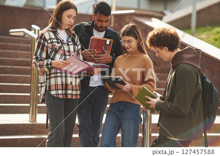With education material in hands, notepads. Four young students in casual clothes are together outdoors 127457588