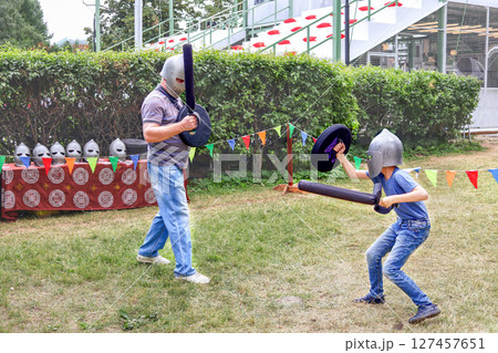 Father and child wearing helmets engage in a playful medieval sword fight with foam swords and shields 127457651