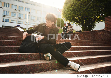 Guy is sitting on the stairs. Young students in casual clothes are together outdoors Guy is sitting on the stairs. Young students in casual clothes are together outdoors 127457684