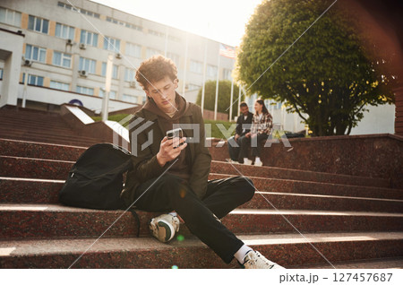 Guy is sitting on the stairs. Young students in casual clothes are together outdoors Guy is sitting on the stairs. Young students in casual clothes are together outdoors 127457687