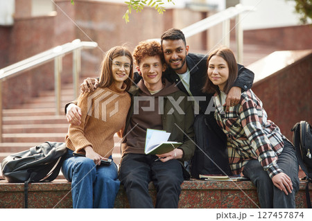 Embracing each other, sitting and smiling. Four young students in casual clothes are together outdoors Embracing each other, sitting and smiling. Four young students in casual clothes are together outdoors 127457874