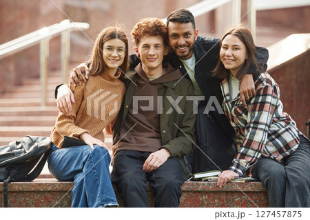 Embracing each other, sitting and smiling. Four young students in casual clothes are together outdoors 127457875