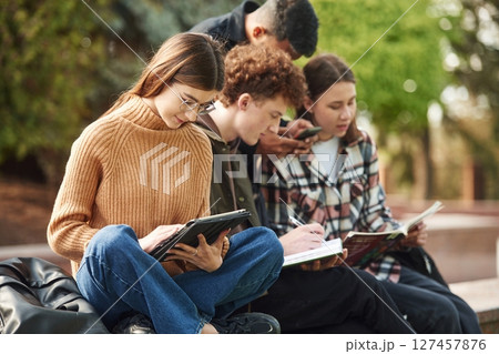 Sitting, reading educational material. Four young students in casual clothes are together outdoors 127457876