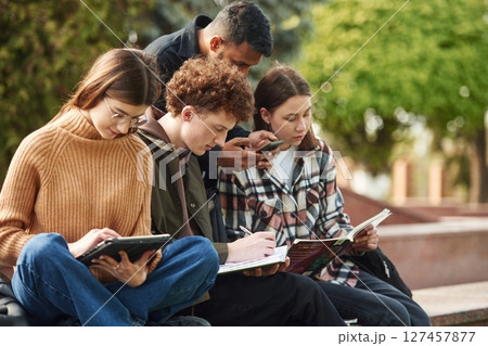 Sitting, reading educational material. Four young students in casual clothes are together outdoors 127457877