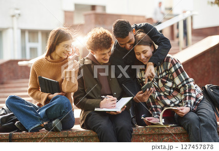 Sitting, reading educational material. Four young students in casual clothes are together outdoors 127457878