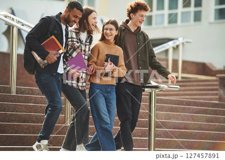 Walking, holding notepads. Four young students in casual clothes are together outdoors Walking, holding notepads. Four young students in casual clothes are together outdoors 127457891