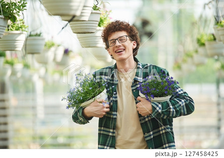 Standing, holding plants in the pots. Young man with curly hair and in glasses is in greenhouse 127458425