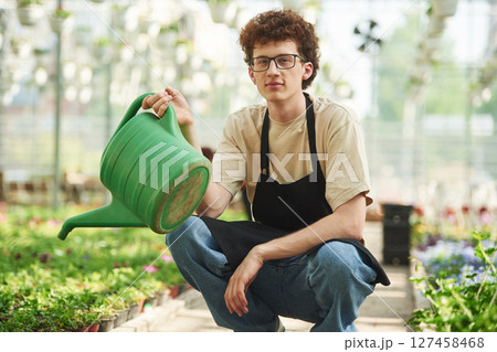 Green watering can in hands. Young man with curly hair and in glasses is in greenhouse 127458468