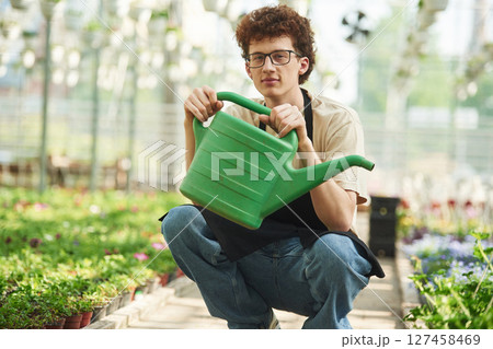 Green watering can in hands. Young man with curly hair and in glasses is in greenhouse Green watering can in hands. Young man with curly hair and in glasses is in greenhouse 127458469
