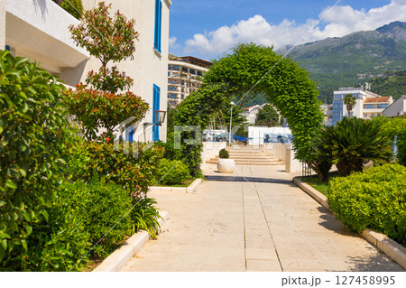 Green arch and mountain view in sunny Mediterranean town. Urban landscaping, nature integration, and scenic architecture with blue shutter details. Green arch and mountain view in sunny Mediterranean town. Urban landscaping, nature integration, and scenic architecture with blue shutter details. 127458995
